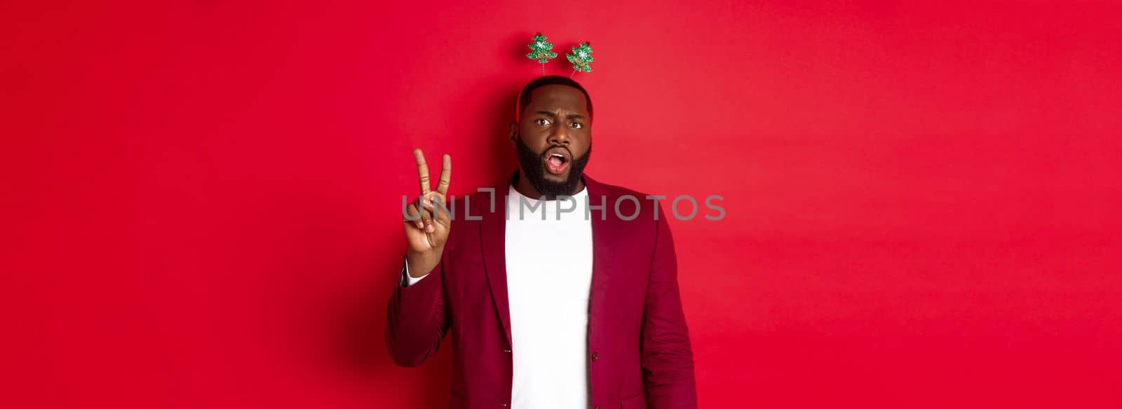 Merry Christmas. Masculine black man in costume and silly party headband, showing peace sign but looking serious, standing over red background.