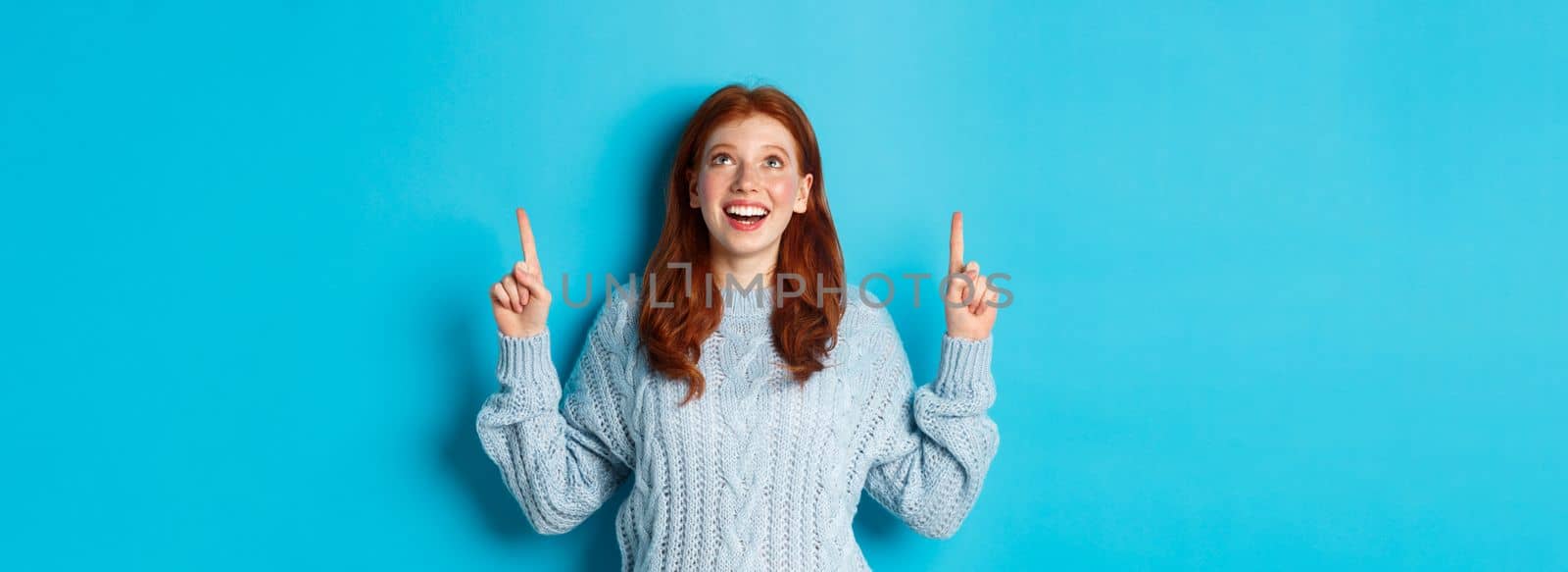 Winter holidays and people concept. Beautiful teenage girl with red hair, wearing sweater, pointing fingers up at logo and smiling, showing advertisement, blue background.