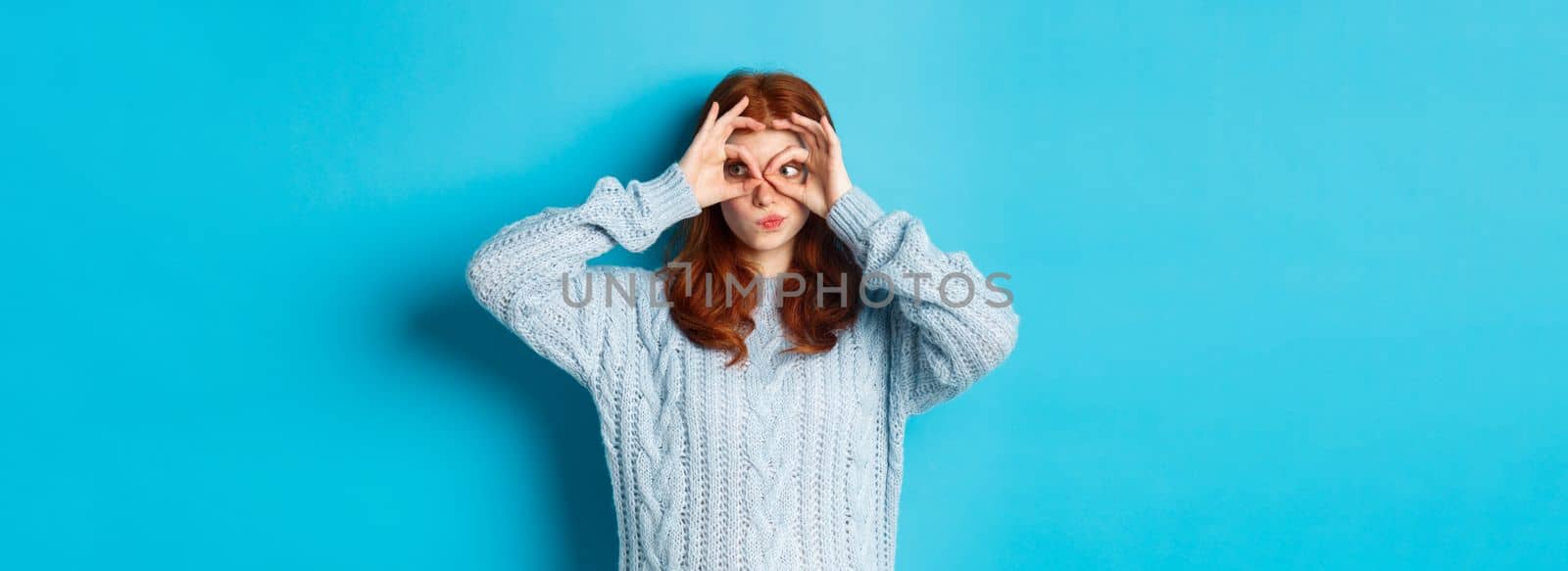 Funny redhead female model in sweater, staring at camera through fingers glasses, express interest and amazement, standing over blue background.