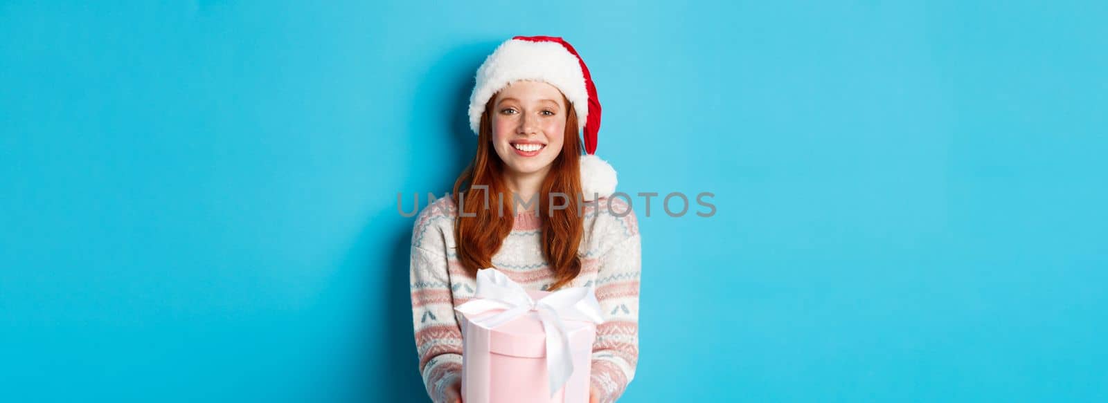 Winter and celebration concept. Beautiful redhead girl in santa hat wishing merry christmas, giving gift and smiling, standing over blue background by Benzoix