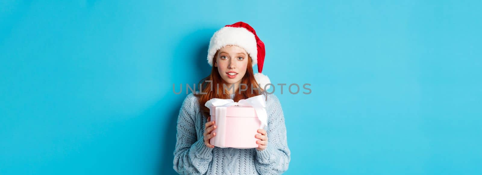 Winter holidays and Christmas Eve concept. Cute redhead girl wearing Santa hat, holding New Year gift and looking at camera, standing against blue background.