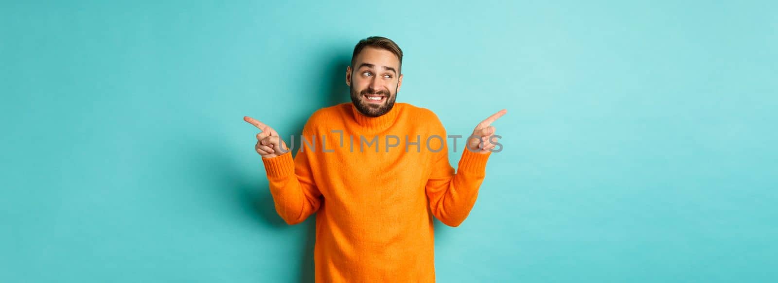 Confused handsome man shrugging indecisive, pointing fingers sideways, cant choose between two variants, standing against light blue background.