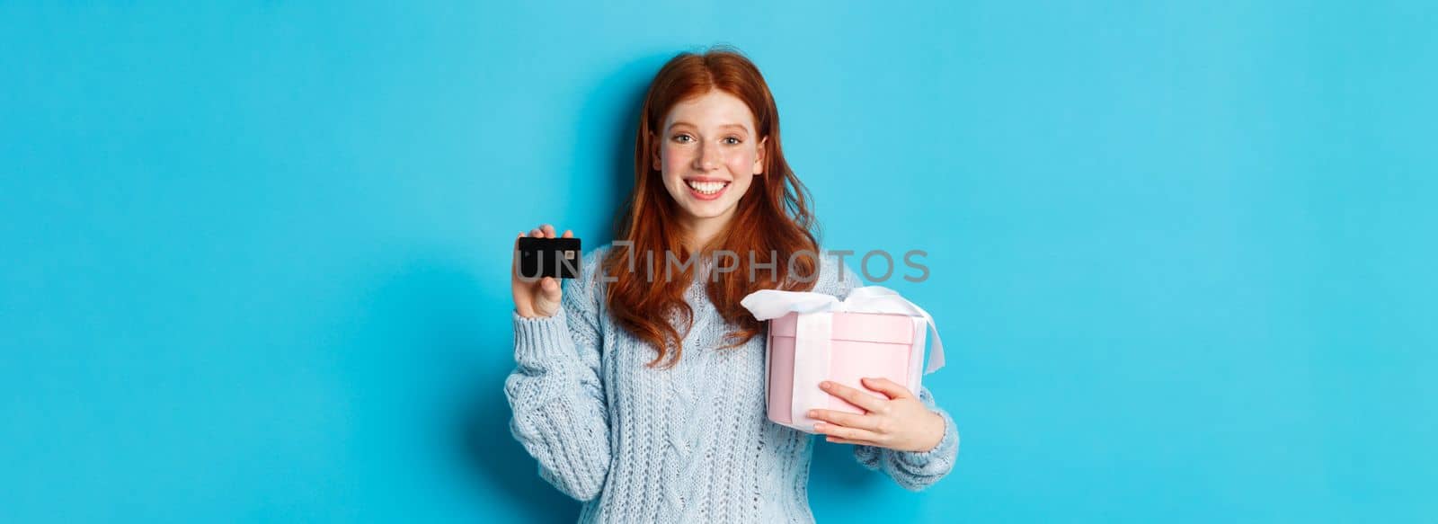 Happy redhead girl buying gifts with credit card, holding box with present and smiling, standing over blue background.