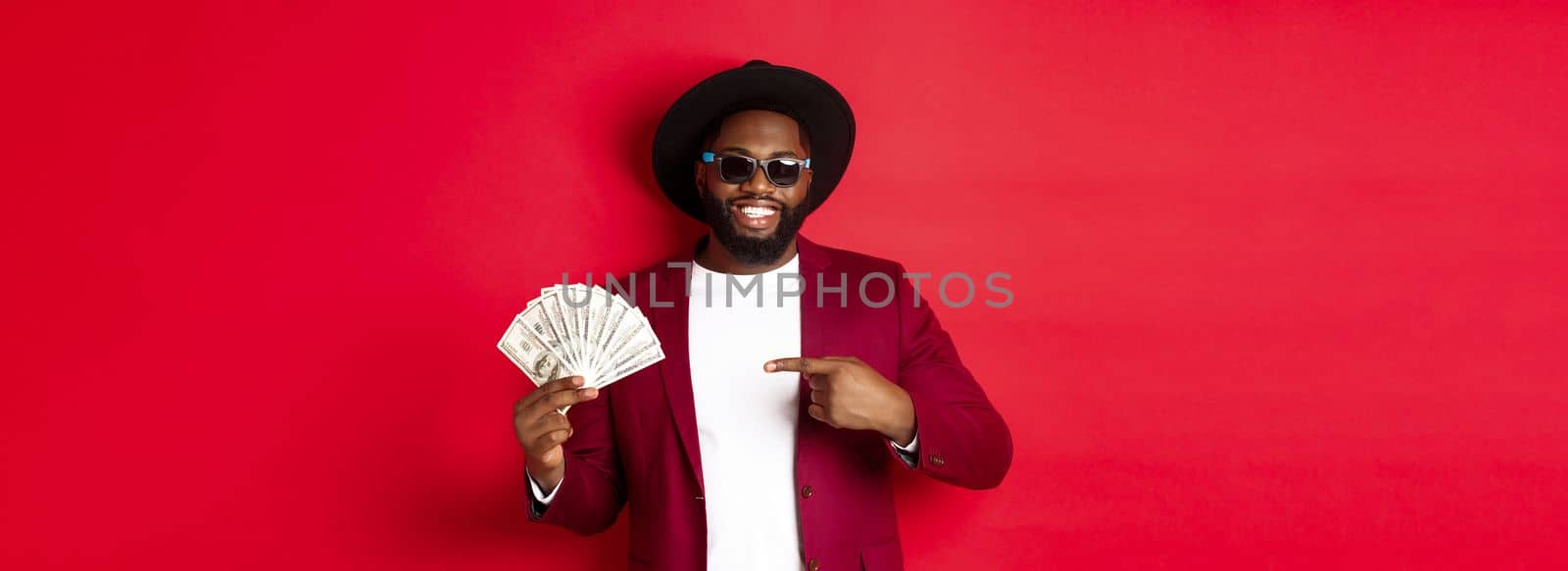 Handsome and stylish african american male model showing money and smiling, wearing sunglasses and fancy hat, standing over red background.