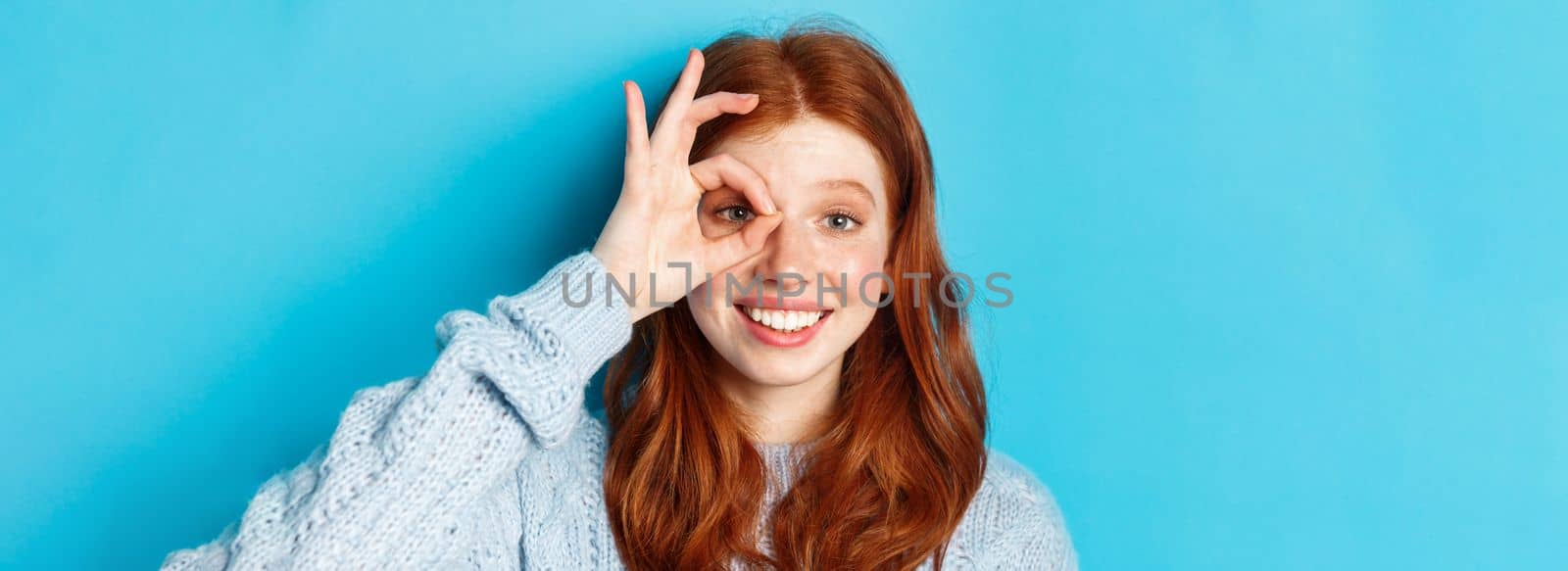 Headshot of cute teenage redhead girl showing okay sign on eye and smiling, standing happy against blue background.