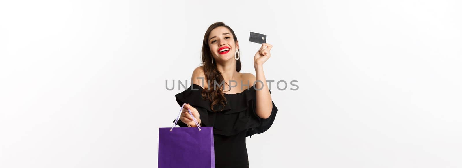 Stylish young woman in black dress going shopping, holding bag and credit card, smiling pleased, standing over white background by Benzoix