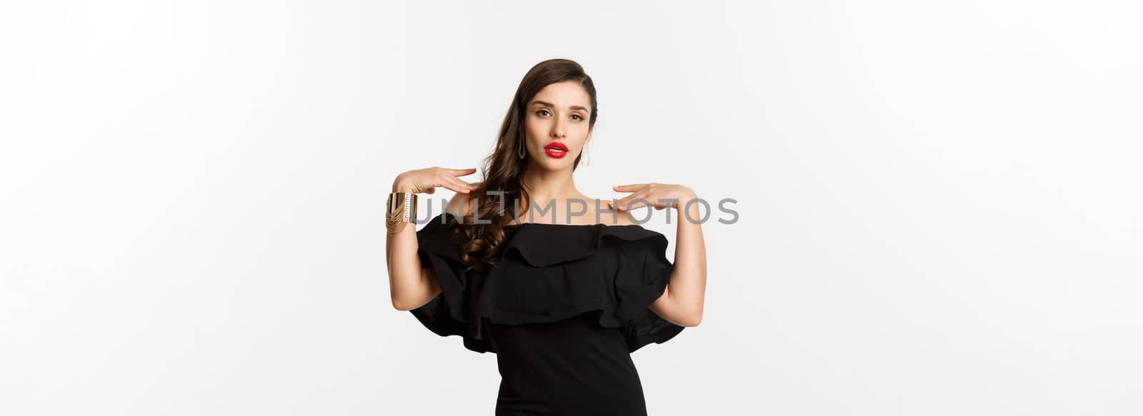 Confident and stylish woman in black elegant dress, looking sassy at camera, standing over white background.