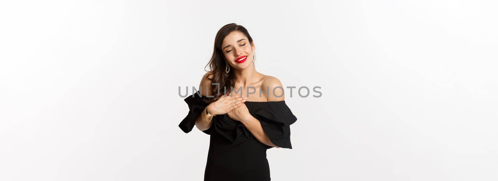 Fashion and beauty. Sensual young woman looking happy, close eyes and holding hands on heart with nostalgic emotion, standing over white background.