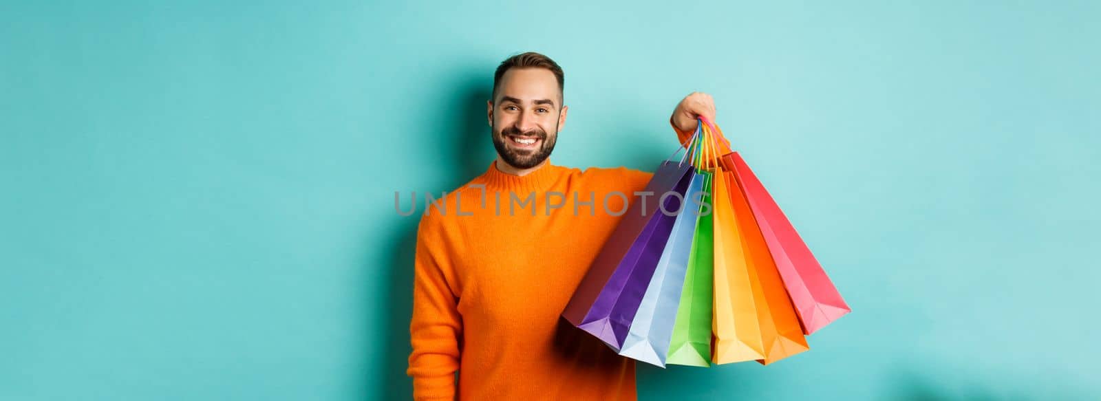 Happy handsome man holding shopping bags and smiling, buying presents, standing over turquoise background by Benzoix