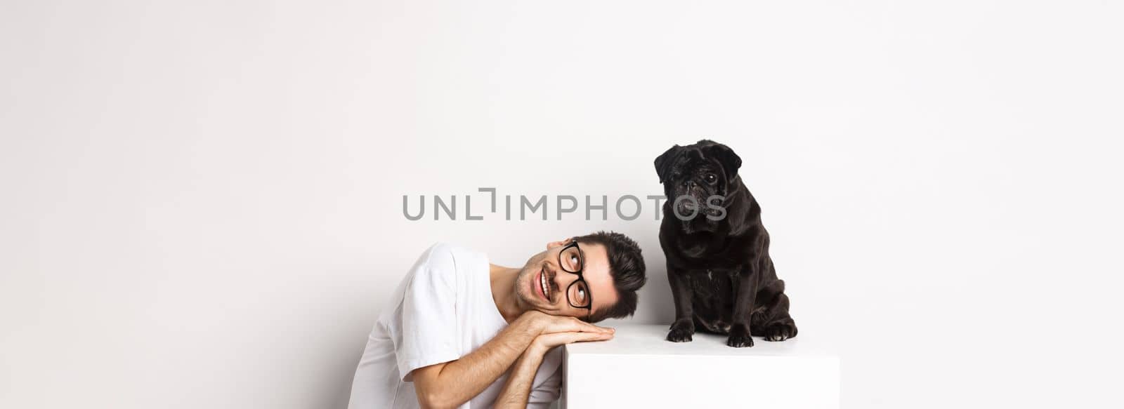 Handsome young man lay head near cute black pug, smiling and looking up at copy space, white background.