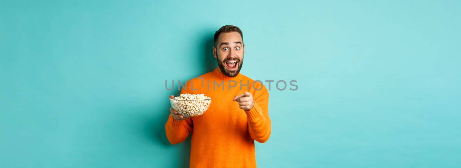 Handsome young man holding bowl of popcorn, smiling amazed and pointing at camera, standing over blue background by Benzoix