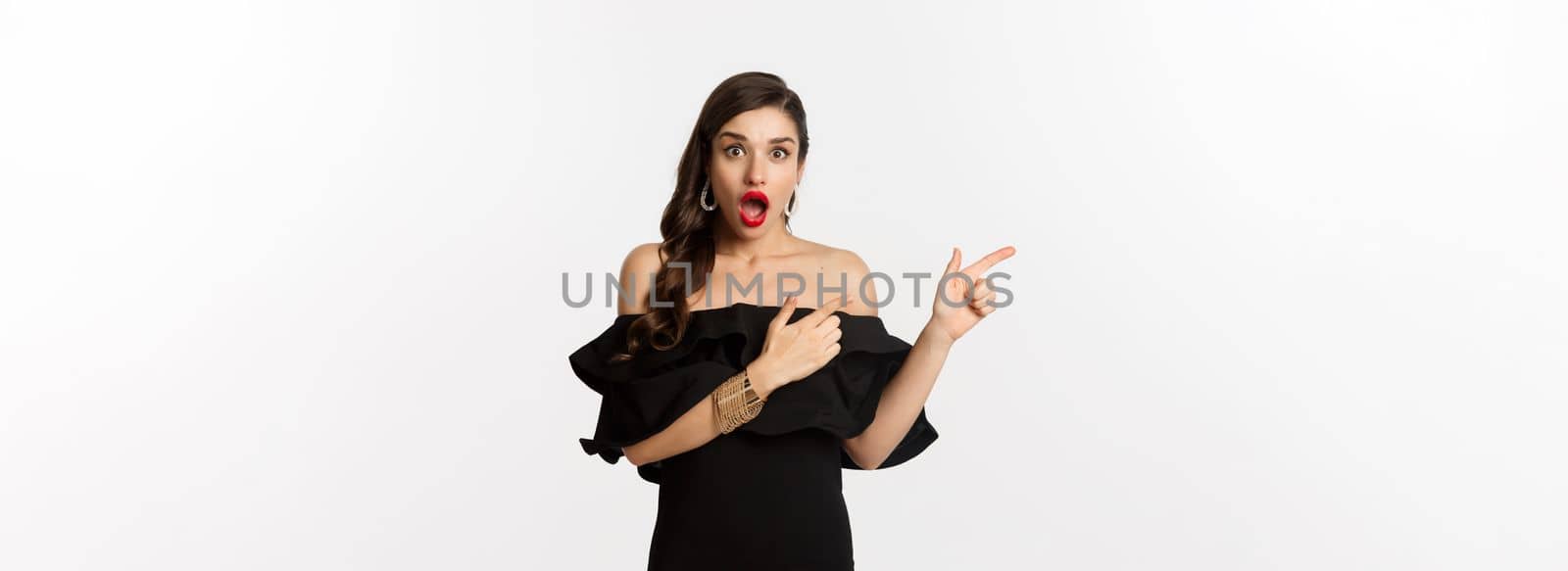 Fashion and beauty. Surprised woman in black glamour dress pointing fingers left, showing advertisement and staring amazed, white background by Benzoix