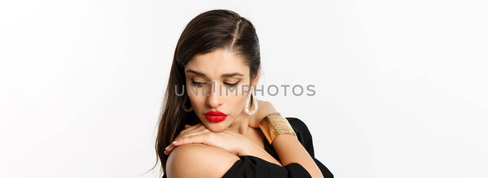 Fashion and beauty concept. Close-up of sensual woman in elegant earrings and black dress, wearing makeup with red lips, looking down tenderly, standing over white background.