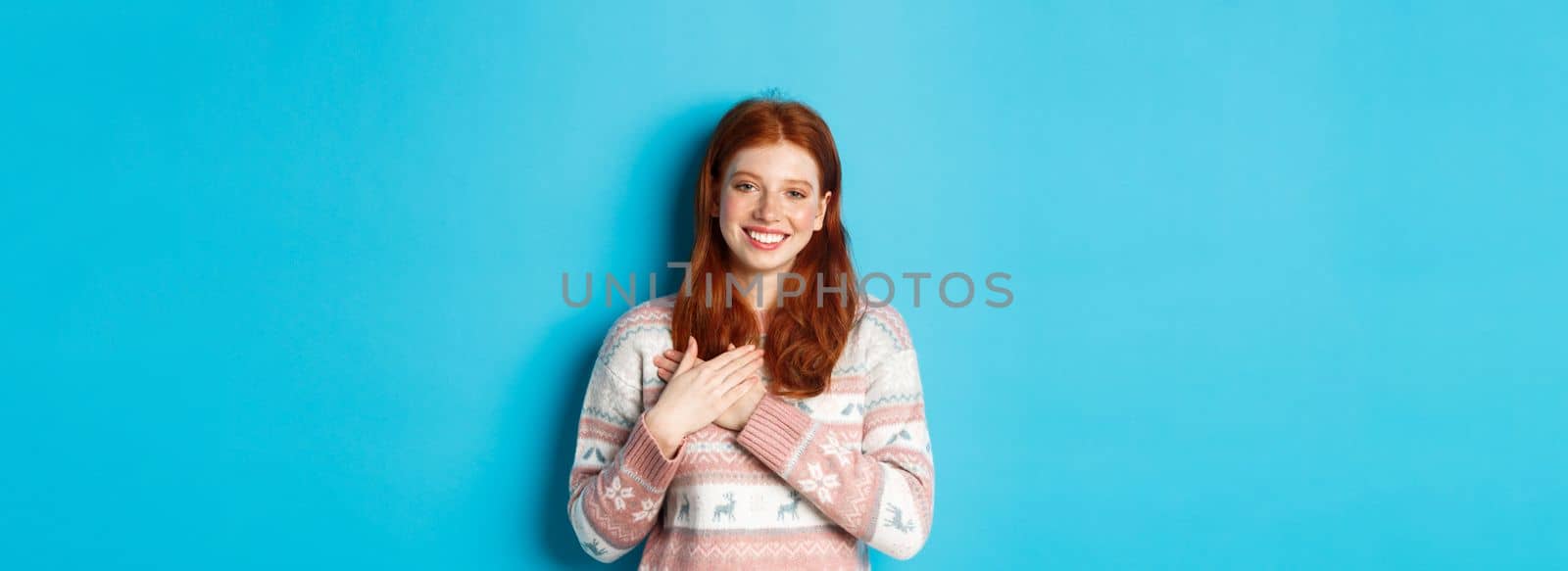 Image of beautiful redhead female model holding hands on heart and smiling, saying thank you, being grateful, standing over blue background.