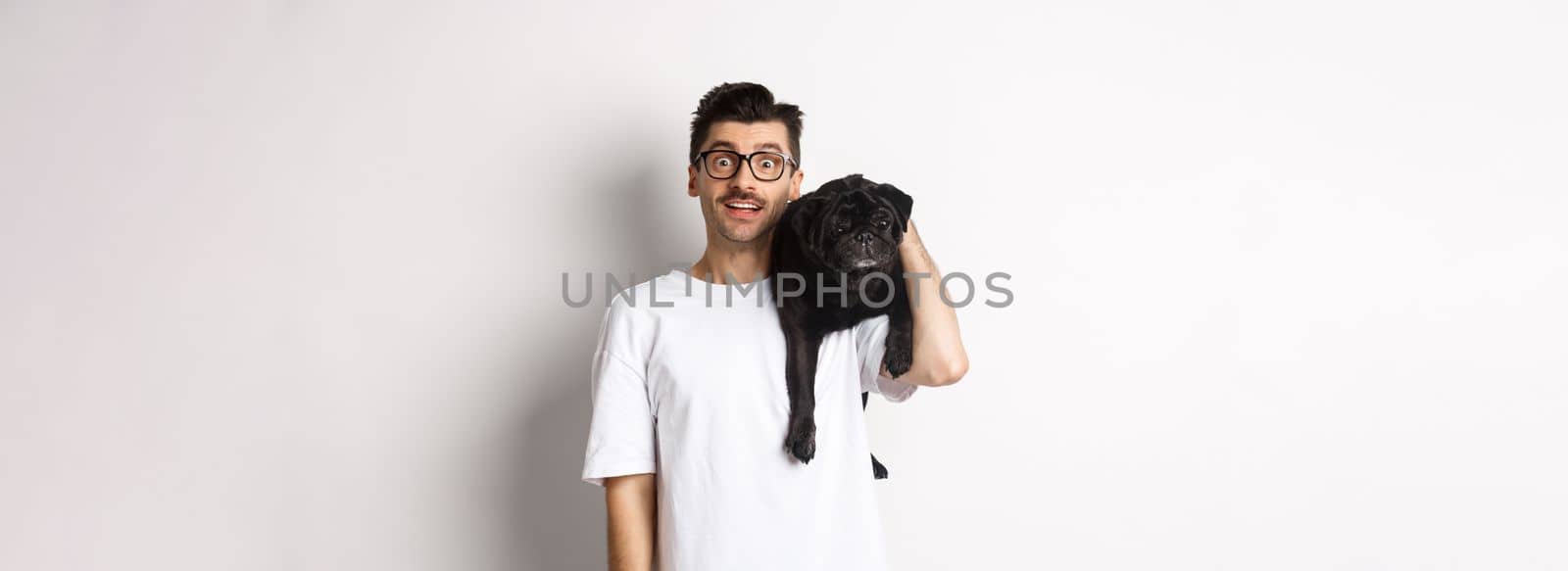 Amazed young man in glasses holding black pug on shoulder and staring at camera impressed. Dog owner posing with cute puppy near white background by Benzoix