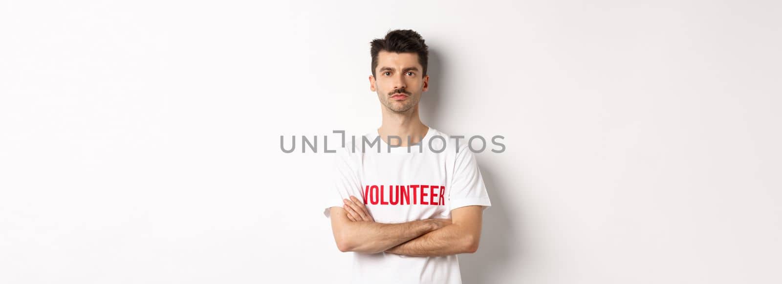 Serious young male volunteer in white t-shirt, holding arms crossed on chest, looking at camera, ready to help.