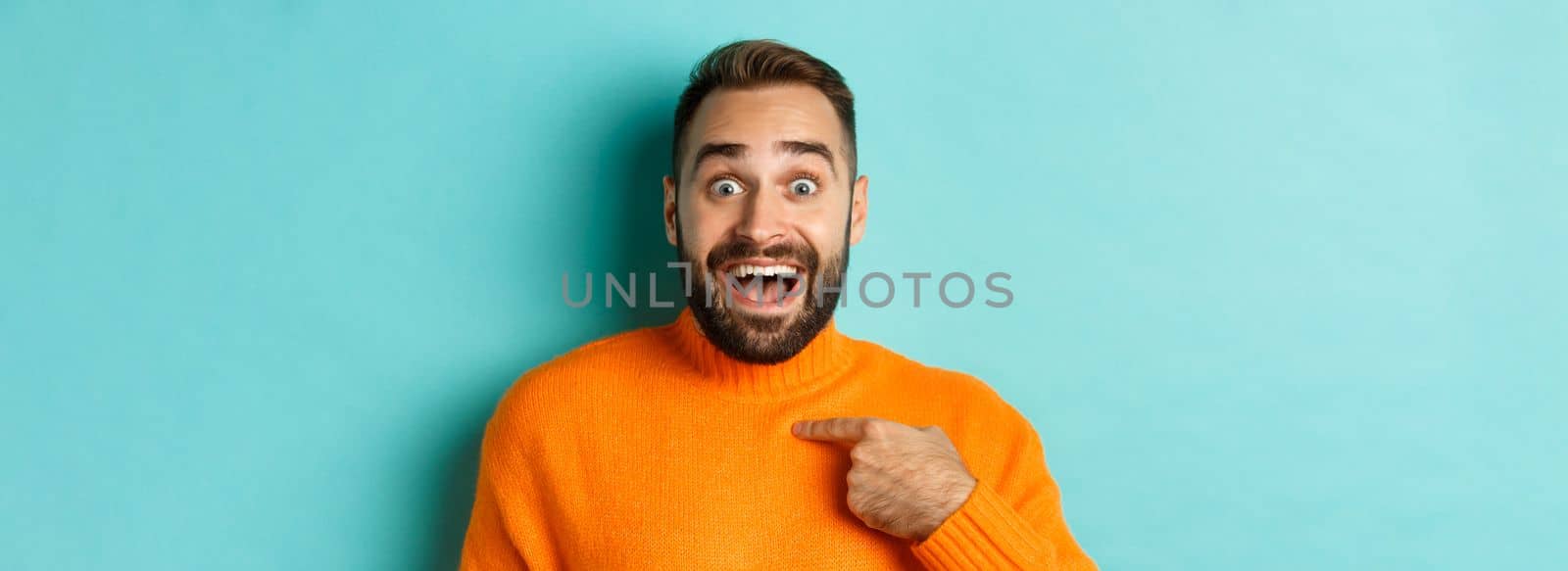 Close-up of happy young man pointing at himself with surprise and excitement, standing over light blue background.