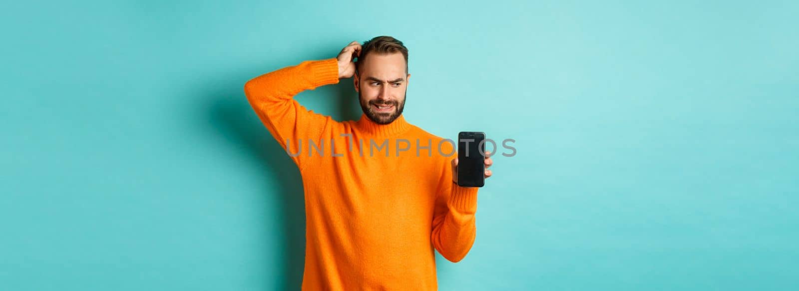 Confused guy scartching head and showing smartphone screen, looking puzzled, standing over turquoise background by Benzoix