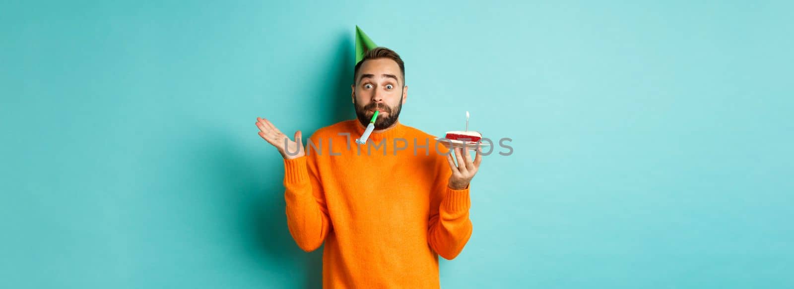 Happy birthday guy celebrating, wearing party hat, blowing wistle and holding bday cake, standing against white background.