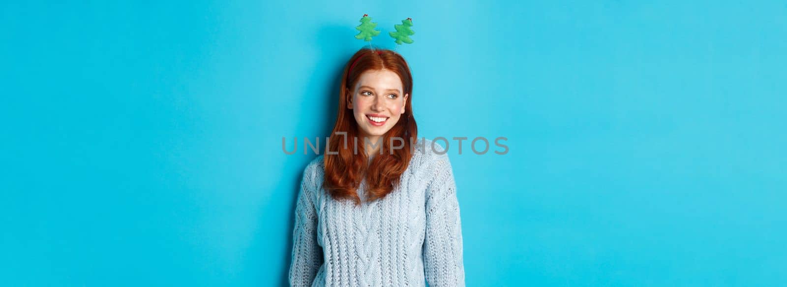 Winter holidays and Christmas sales concept. Cute redhead girl in funny New Year headband smiling, looking left at logo, standing over blue background.