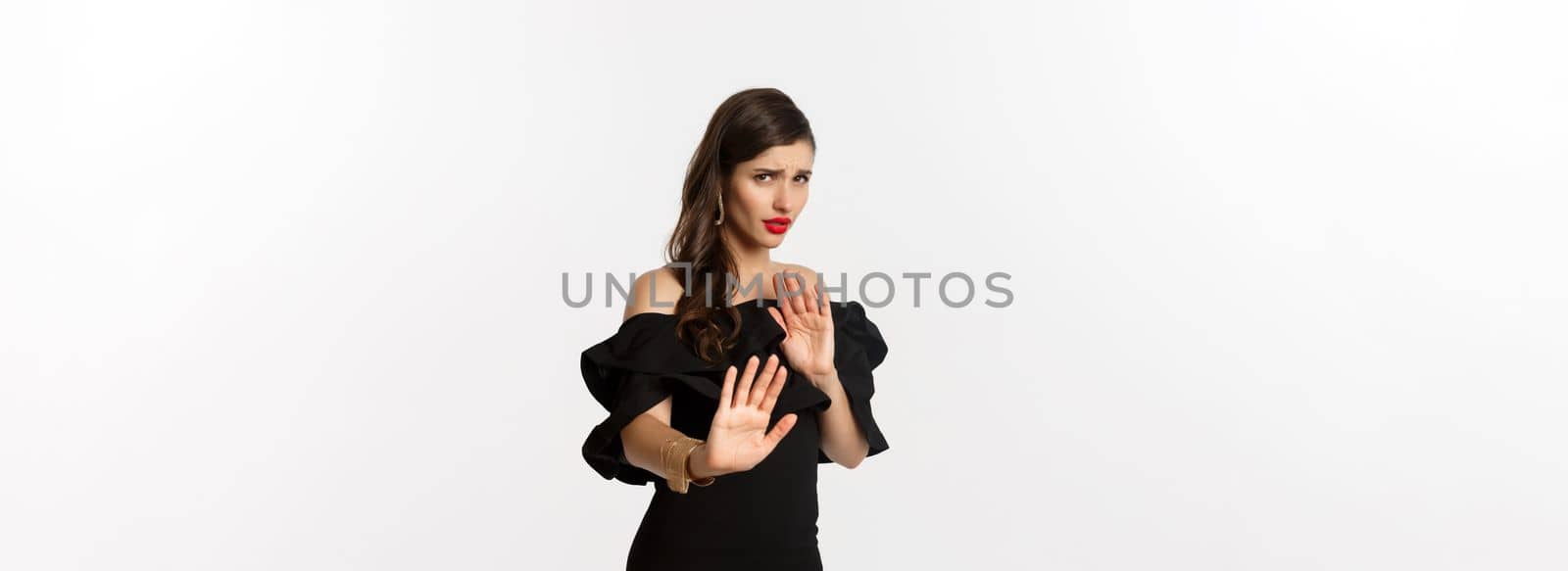 Fashion and beauty. Reluctant and worried woman asking to stay away, showing stop gesture and looking scared, standing in black dress over white background by Benzoix