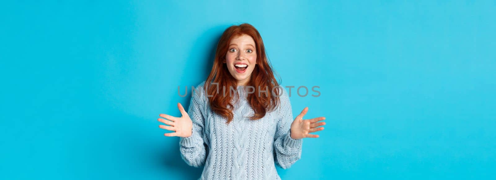 Excited redhead woman shaking hands, explaining big news, staring at camera amazed, standing against blue background.