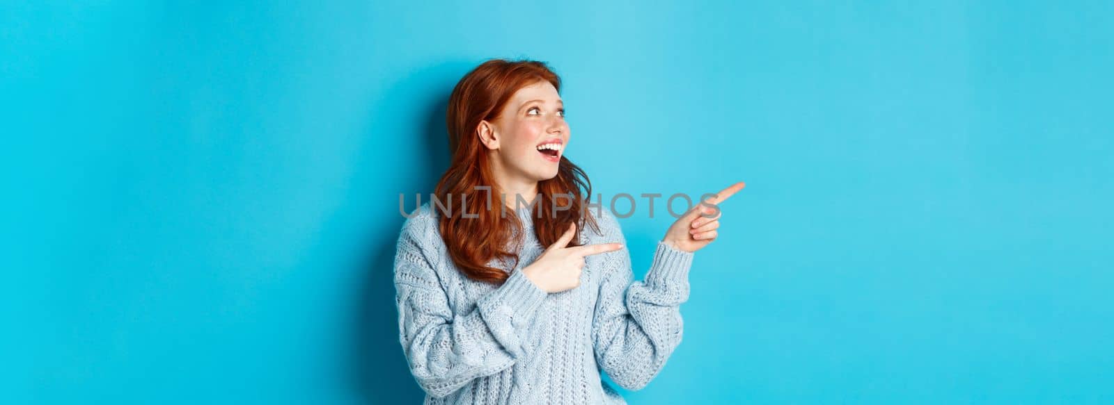 Excited redhead girl in sweater, looking and pointing fingers left, showing promo offer or logo, standing over blue background.