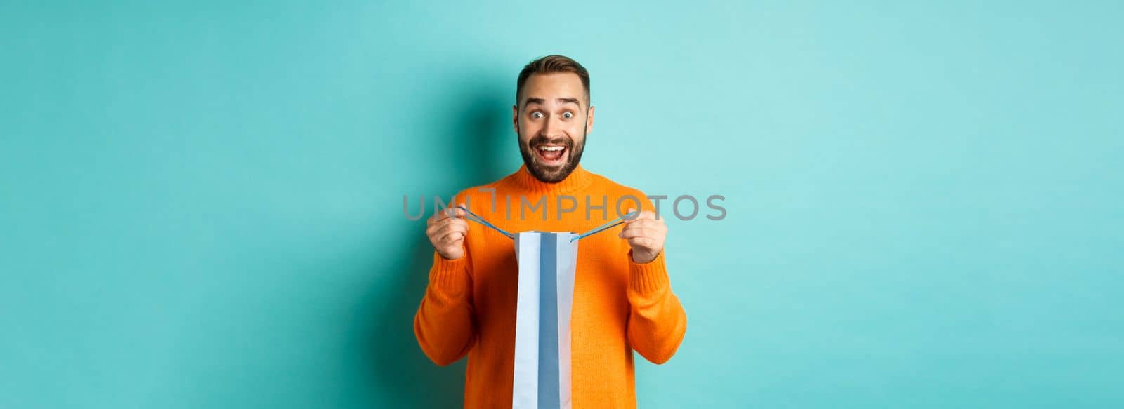 Surprised man open shopping bag and looking amazed, receiving gift on holiday, standing over turquoise background.
