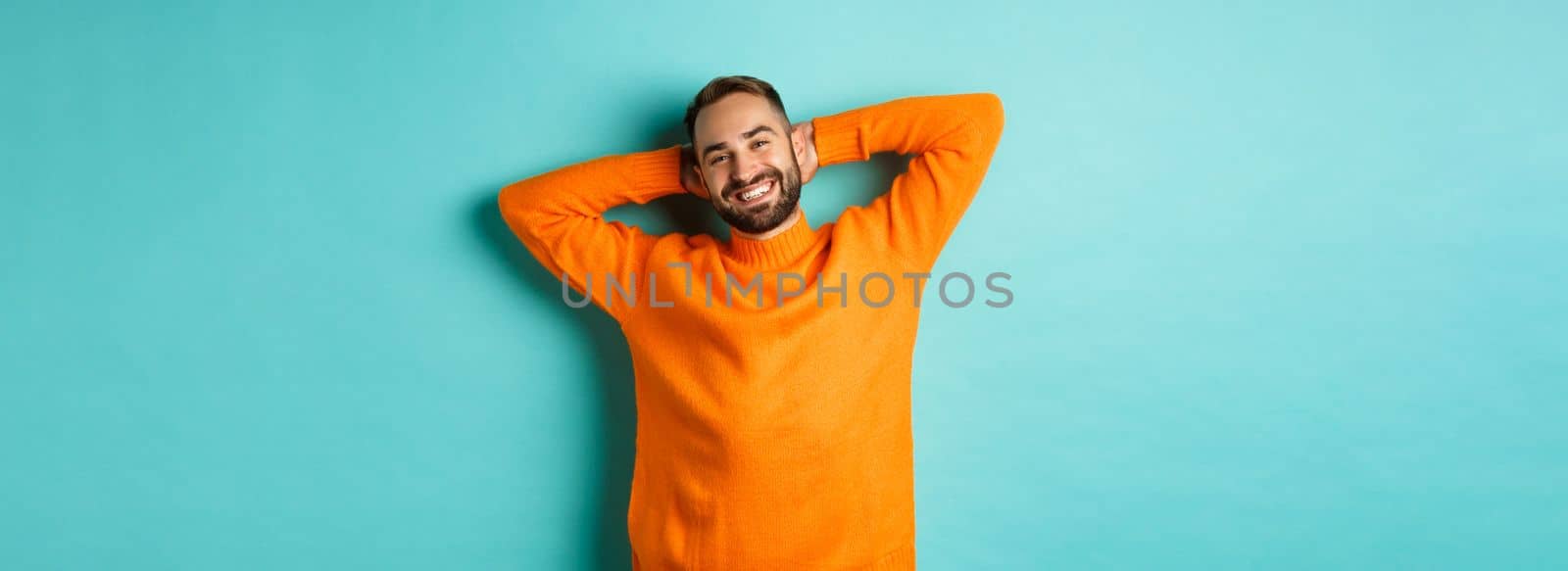 Handsome man relaxing, resting and smiling, holding hands behind head with carefree face, standing against light blue background.