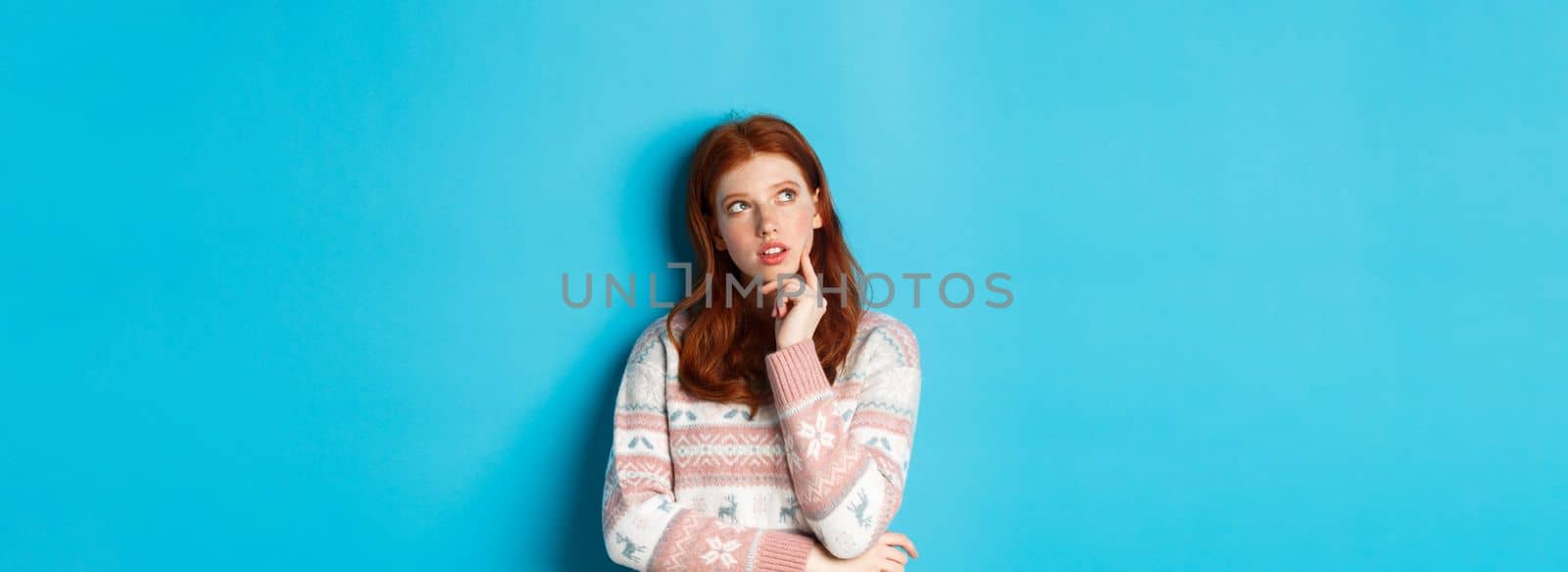 Image of thoughtful redhead girl looking at upper left corner, thinking or making choice, standing in winter sweater over blue background.