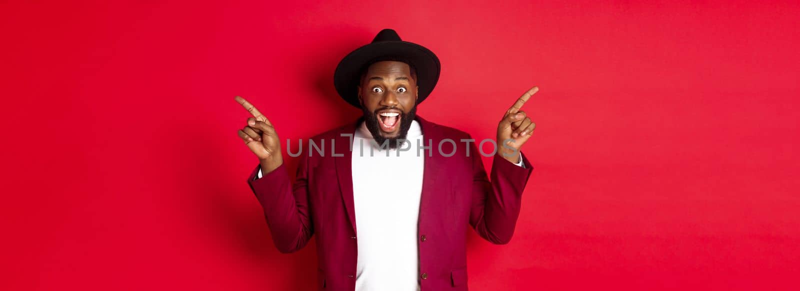 Winter holidays and shopping concept. Cheerful Black man smiling and showing two promo, pointing fingers sideways at copy spaces, red background.