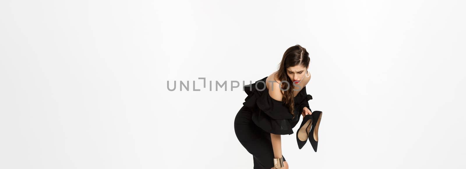 Beauty and fashion concept. Full length of woman feeling pain in feet, take-off high heels and rubbing foot with tired face, standing in black dress over white background.