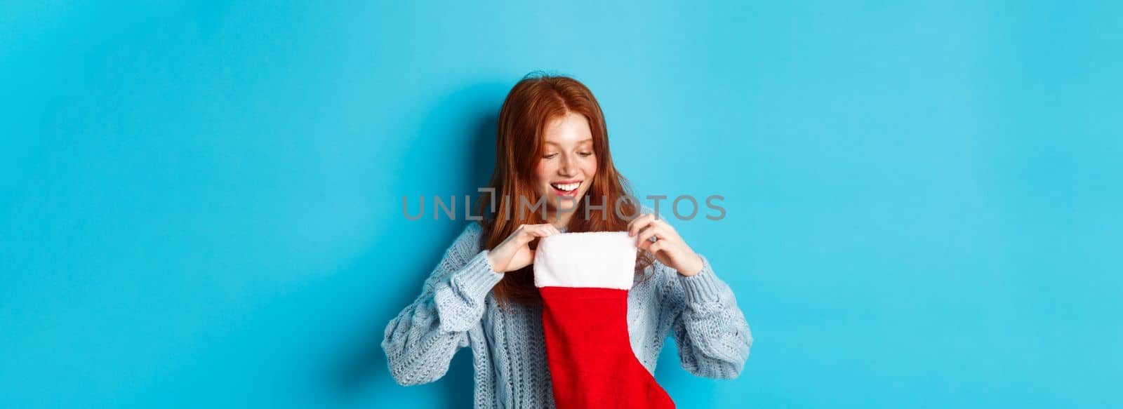 Winter holidays and gifts concept. Funny redhead girl looking inside Christmas stocking and smiling happy, receiving xmas present, standing against blue background.