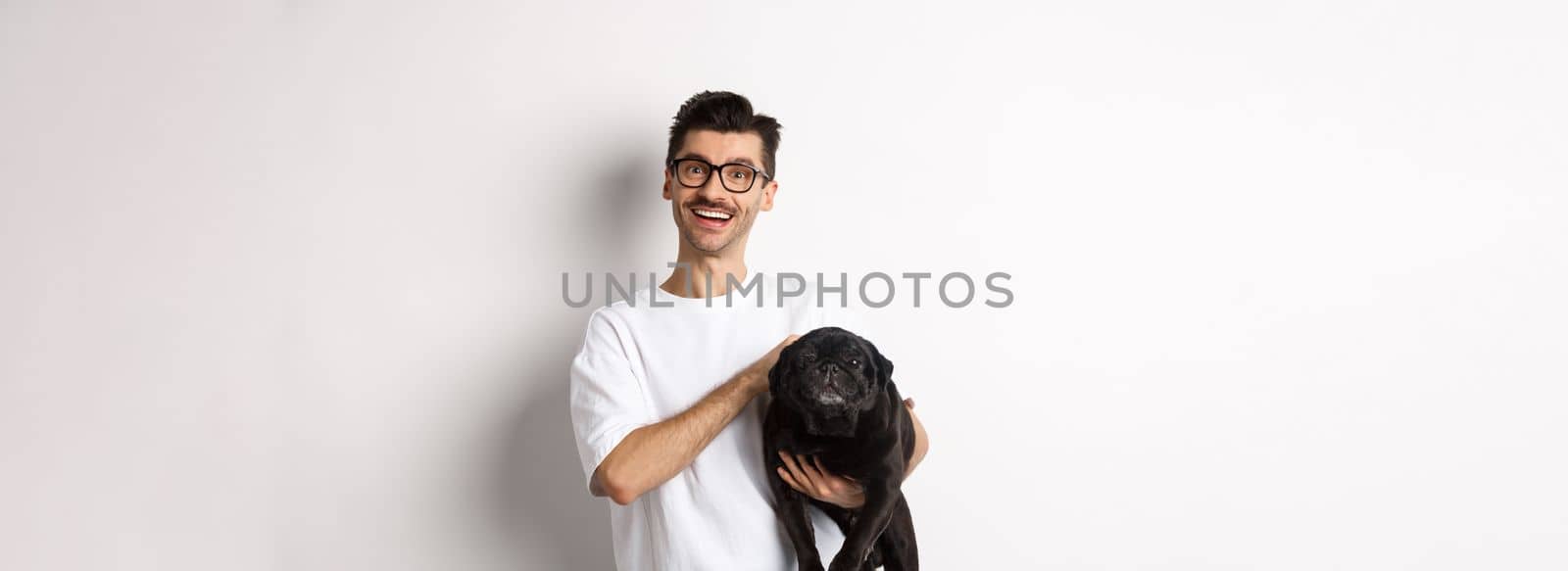 Happy hipster guy in glasses pet dog and smiling. Cute black pug enjoy spending time with owner, looking satisfied, standing over white background by Benzoix