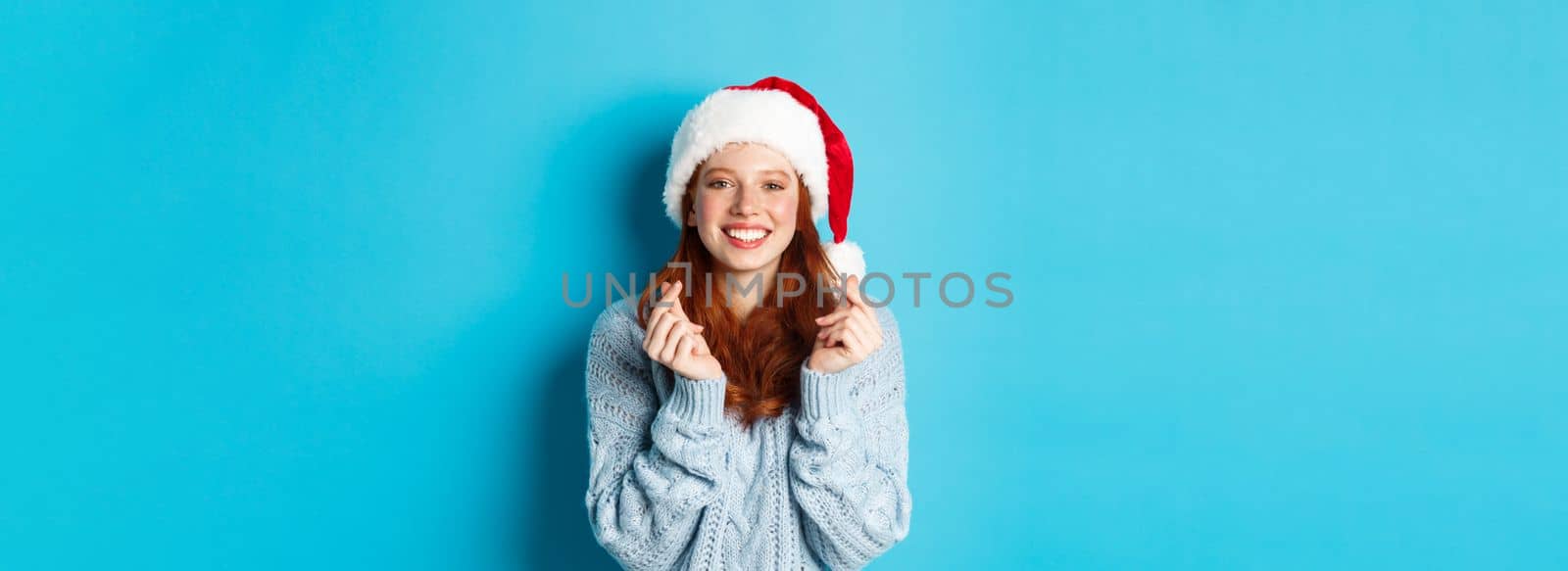 Winter holidays and Christmas Eve concept. Hopeful redhead girl in santa hat, making wish on xmas with fingers crossed, wearing santa hat, standing over blue background.