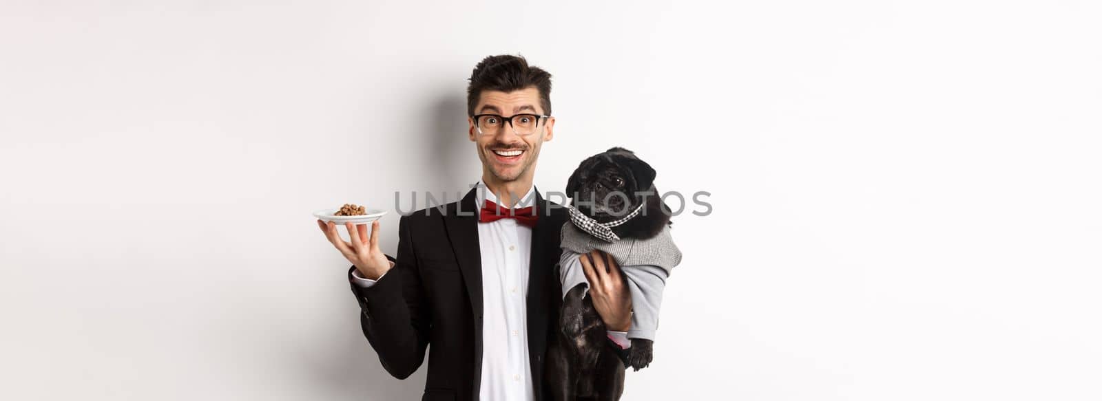 Handsome young hipster in suit and glasses holding cute black pug and pet food on plate, standing over white background by Benzoix