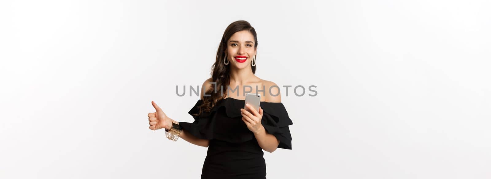 Online shopping concept. Attractive woman in trendy black dress, makeup, showing thumb-up and using mobile phone app, white background by Benzoix