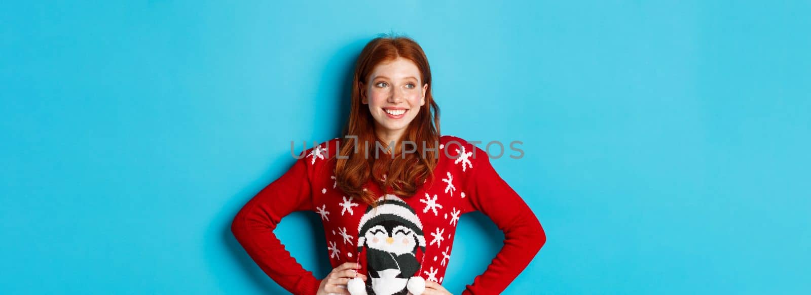 Winter holidays and Christmas Eve concept. Beautiful teenage redhead girl in xmas sweater looking left at logo, smiling pleased, holding hands on waist, blue background by Benzoix