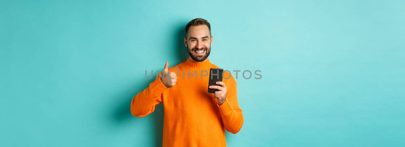 Photo of satisfied young man in orange sweater, showing thumb-up after reading on mobile phone, standing pleased against turquoise background.