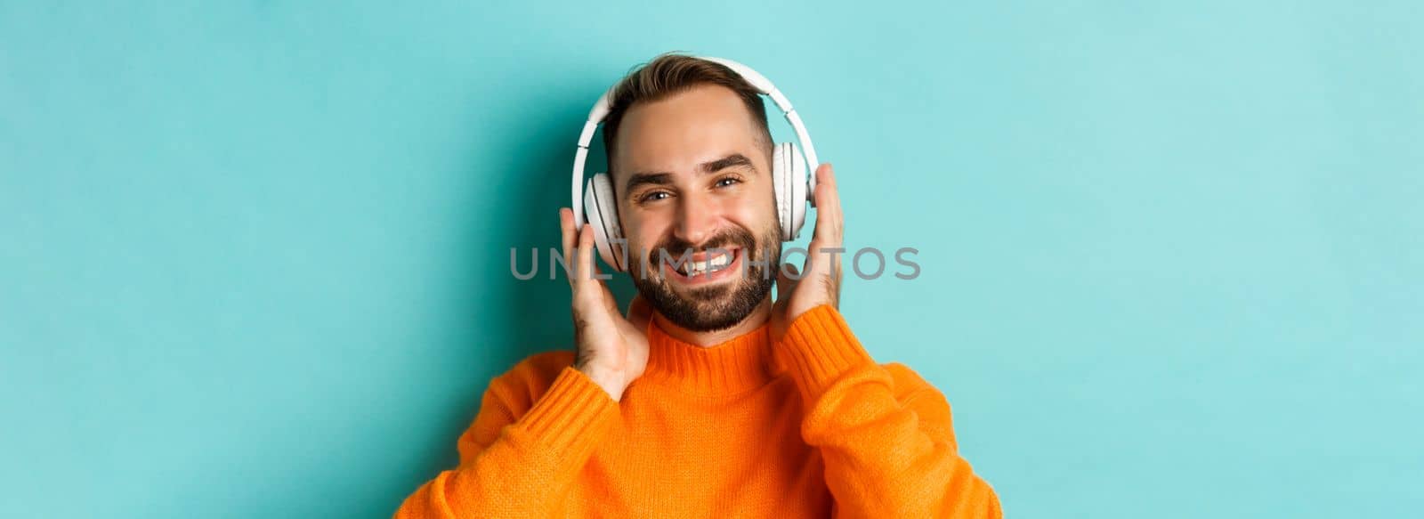 Close-up of handsome modern man listening music in headphones, standing in orange sweater over turquoise background by Benzoix
