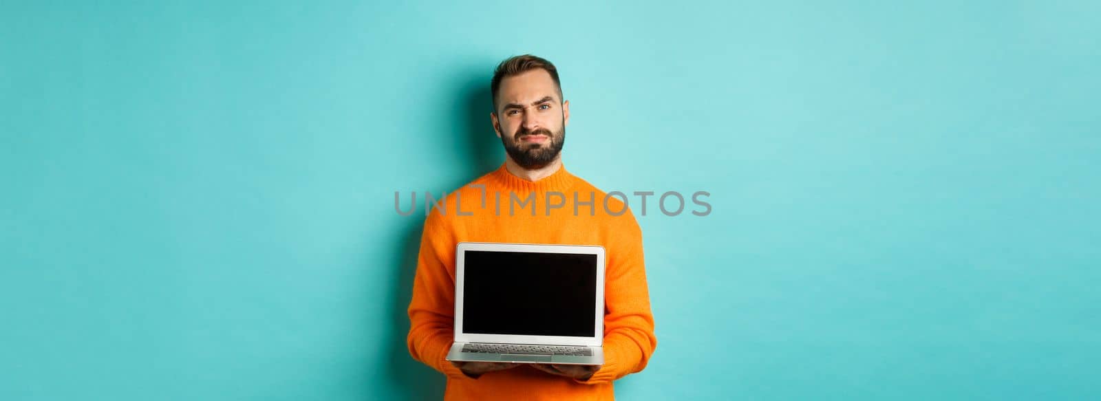 Handsome bearded man in orange sweater showing laptop screen, demonstrating promo, grimacing disappointed and upset standing over light blue background by Benzoix