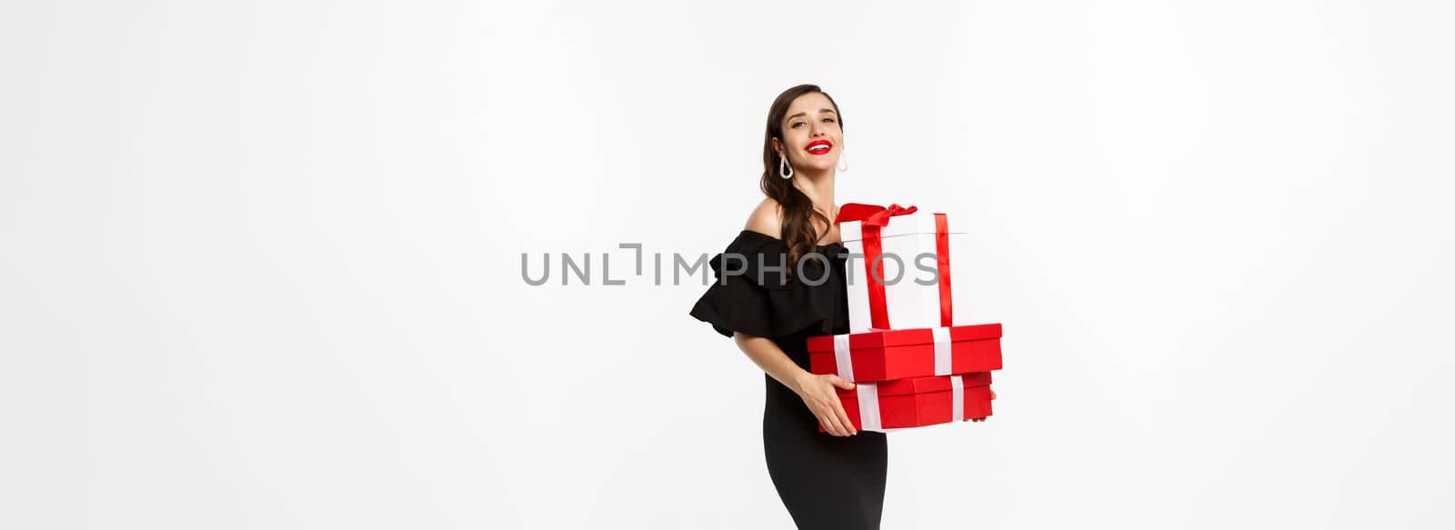 Full length of elegant woman in black dress, red lips, holding christmas presents and smiling pleased, receive gifts, standing over white background.
