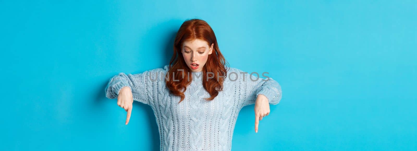 Winter holidays and people concept. Impressed redhead girl in sweater, looking and pointing down with amazement, standing against blue background.