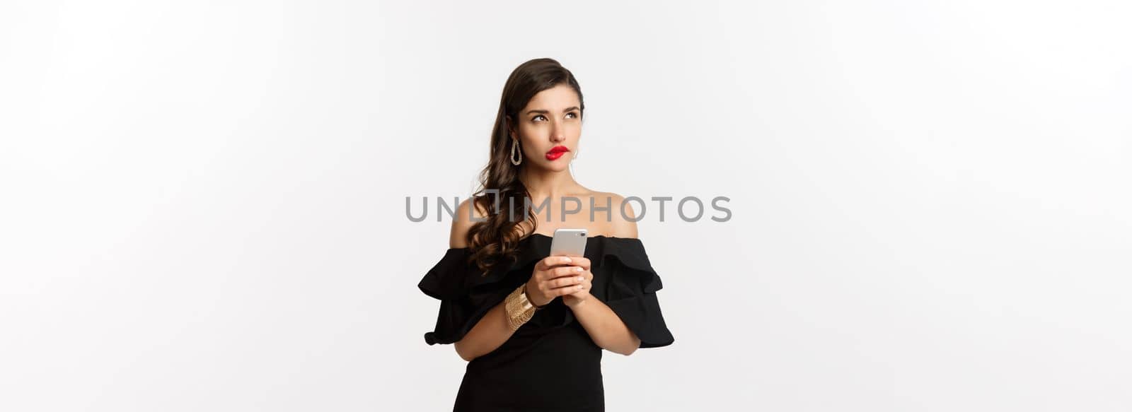 Thoughtful young woman using smartphone and thinking, looking up and pondering, standing over white background. Copy space