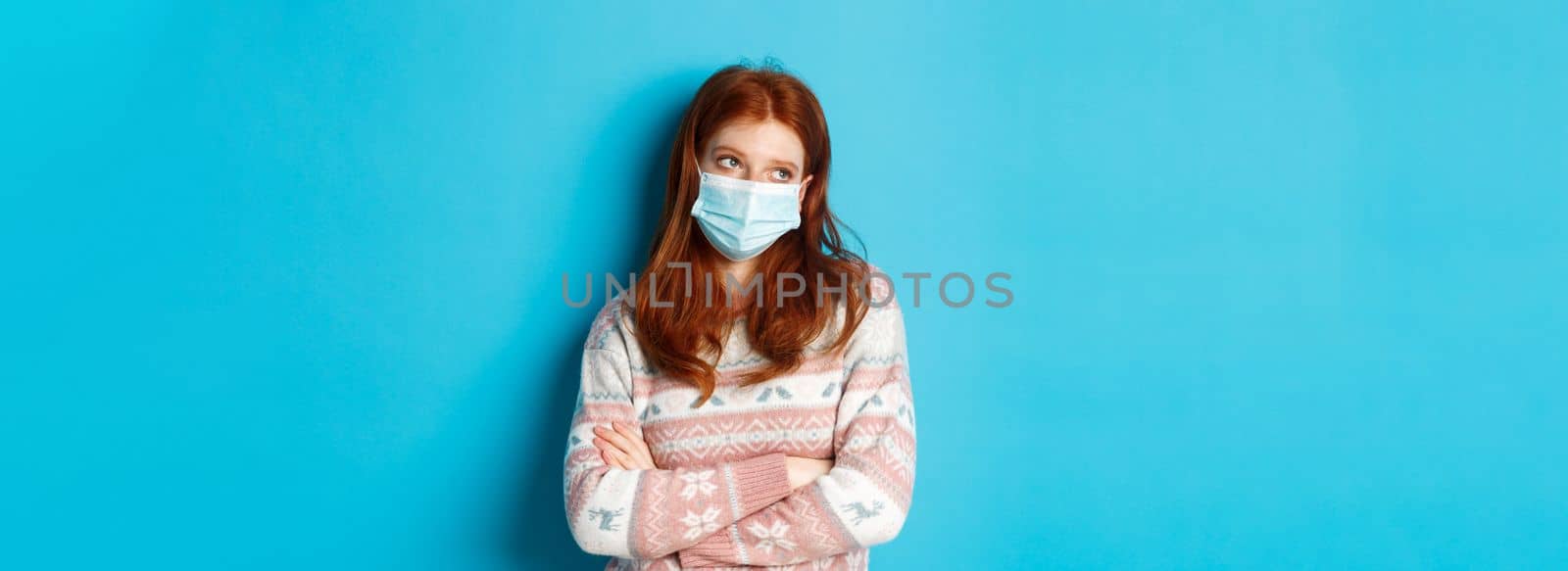 Winter, covid-19 and pandemic concept. Ignorant redhead teenage girl in face mask, roll eyes and looking unamused, cross arms on chest reluctant, standing over blue background by Benzoix