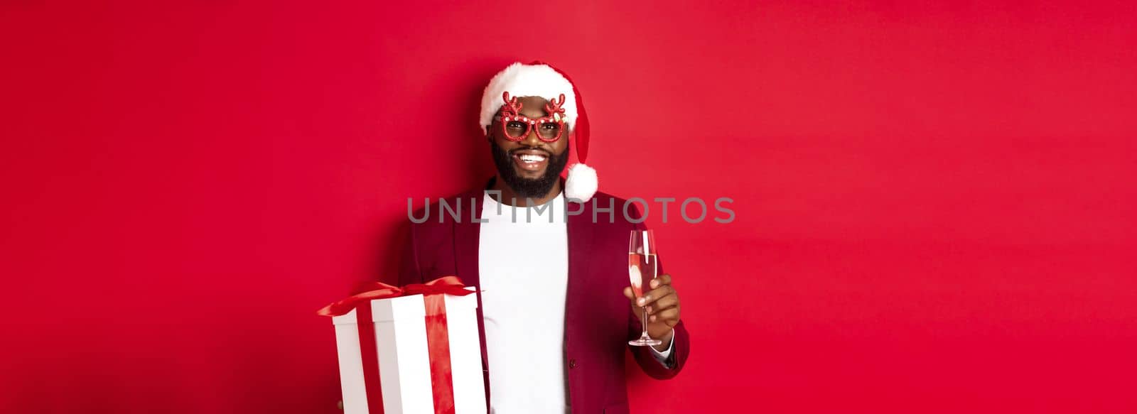 Christmas. Handsome african american man in party glasses and santa hat, holding new year gift and glass of champagne, wishing happy holidays, red background.