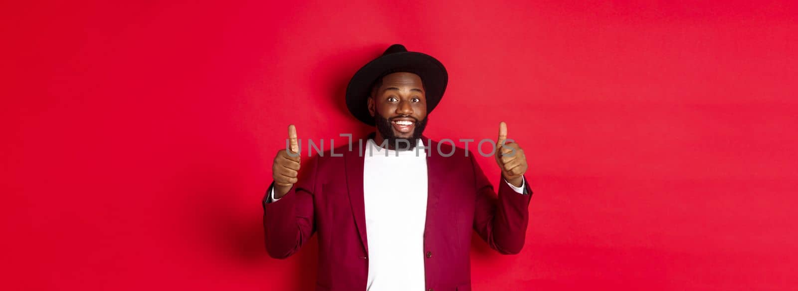 Fashion and party concept. Cheerful african american man in classy jacket, celebrating New Year xmas, showing thumbs up and smiling, like and approve, red background.