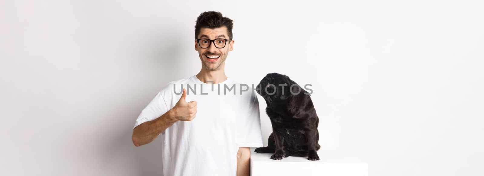 Cheerful handsome guy standing near cute black pug and showing thumb-up. Pet owner approve and recommend product for dogs, white background.