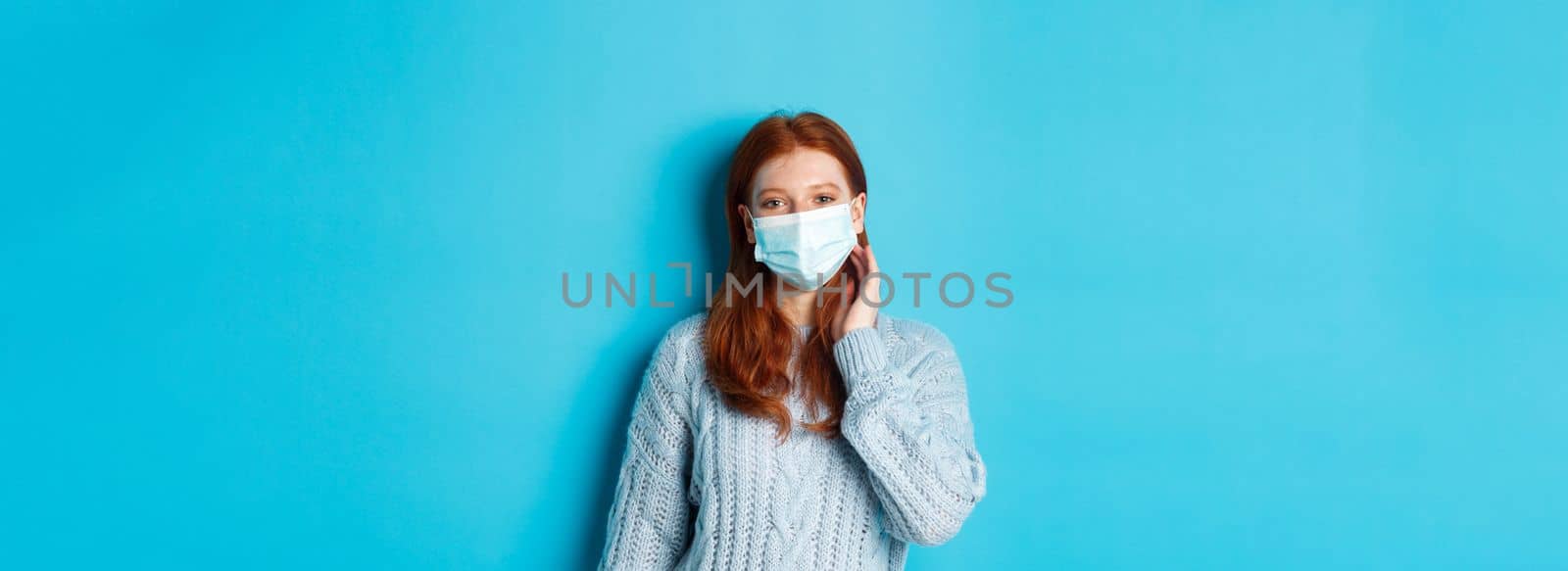 Winter, covid-19 and social distancing concept. Beautiful redhead teenage girl, wearing face mask and tuck hair strand behind ear, staring at camera, standing over blue background.