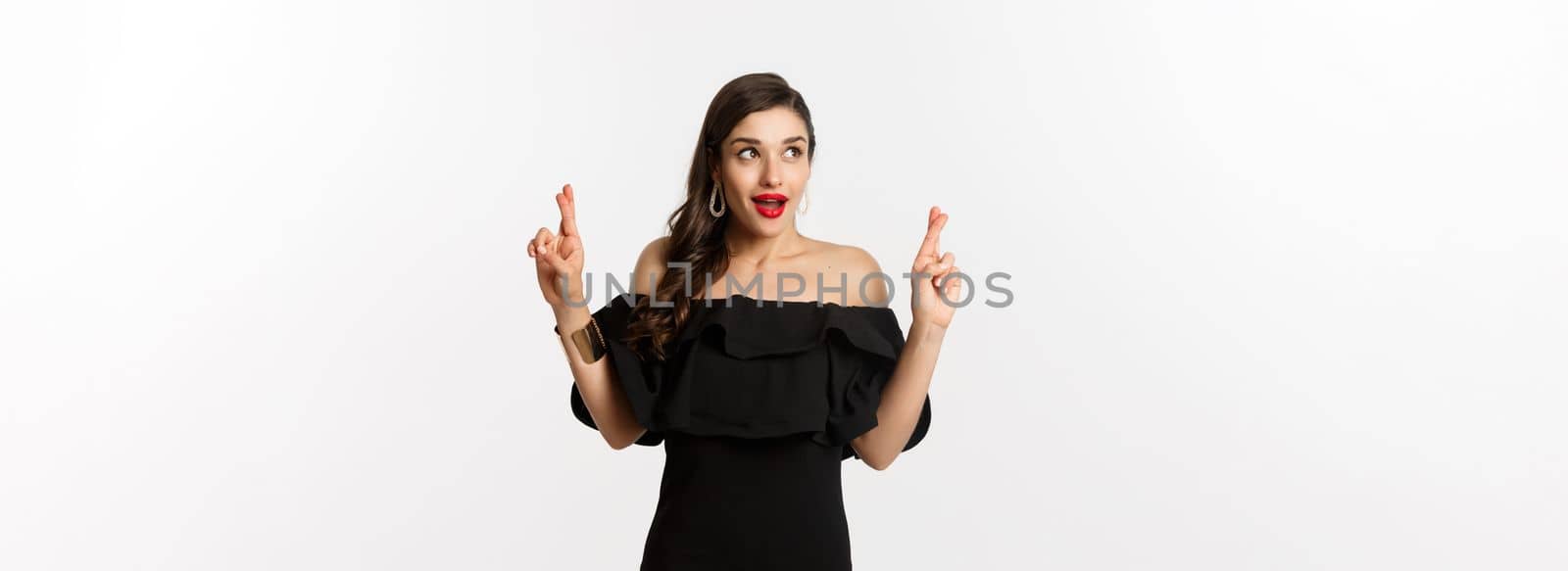 Fashion and beauty. Excited gorgeous woman in black dress, holding fingers crossed and looking at upper left corner, making wish, standing over white background.